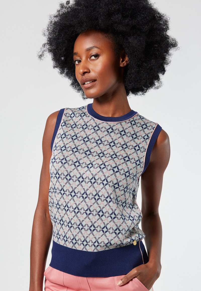 Sleeveless knit sweater features a geometric pattern in pale pink and navy on a light background, with a ribbed navy hem and side button detail.