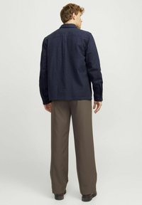 Uomo che indossa una giacca blu scura, pantaloni marroni e scarpe nere, in piedi e di spalle contro uno sfondo chiaro e uniforme.
