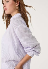 Helllila gestreifte Bluse mit Kragen zum Knöpfen. Weicher, strukturierter Stoff mit hochgekrempelten Ärmeln, entspannter Schnitt und seitlichen Taschen.