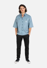 Joven con cabello de longitud media que lleva una camisa azul clara de botones holgada, pantalones negros y zapatillas negras con suelas blancas.