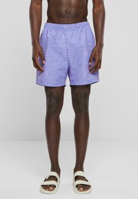 Shorts de bain violets avec un motif subtil, une taille élastique et une coupe droite. Associés à des sandales blanches à bout ouvert.