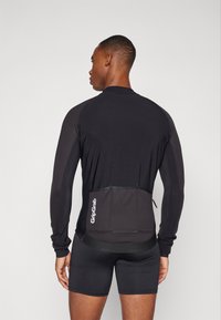 Langärmliges Sportoberteil in Schwarz, mit einer Rückentasche, glattem Stoff und Logodetail am Saum. Getragen mit schwarzen Shorts.
