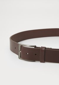 Cinturón de cuero marrón con un acabado suave, que presenta una hebilla de metal plateado y múltiples agujeros de ajuste. Diseño sencillo, material resistente.