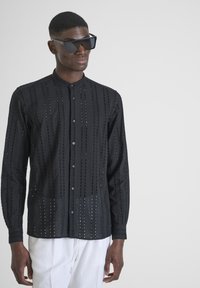 Camicia nera trasparente con modelli a taglio verticale, maniche lunghe e colletto a fascia, abbinata a grandi occhiali da sole neri.