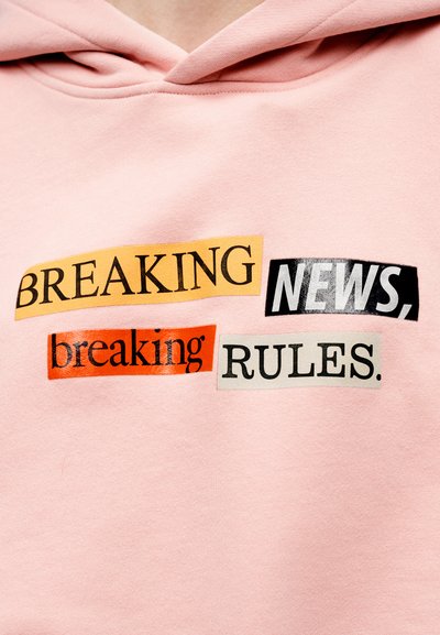 Ροζ φούτερ με κουκούλα με έντονα μπαλώματα κειμένου σε πορτοκαλί, μαύρο και κίτρινο: "BREAKING NEWS", "breaking" και "RULES." Μαλακό ύφασμα, καθημερινός σχεδιασμός.