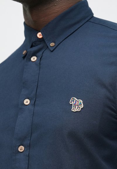 Tmavomodrá košeľa so zapínaním na gombíky vyrobená z látky s hladkým povrchom. Má dekoratívnu výšivku koní v farebných prúžkoch a hnedé gombíky.