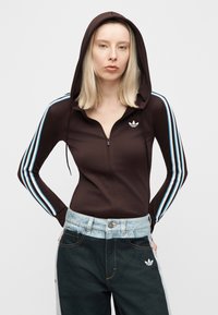 Γυναίκα που φοράει μαύρο φούτερ με φερμουάρ, με μπλε και λευκές ρίγες στα μανίκια και φαρδιά δίχρωμα τζιν με τα χέρια στις τσέπες.