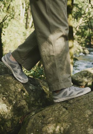 Osoba s sivim tenisicama i maslinastim hlačama koja hoda po mahovinom prekrivenim stijenama u šumi blizu tekuće rijeke.