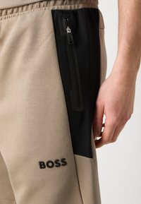 Beige atletske kratke hlače z črnimi mrežastimi žepi, opremljene z zadrgo in vtisnjenim logotipom "BOSS" na levem stegnu.