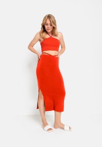 Rotes, geripptes Crop-Top mit Halterdesign, kombiniert mit einem passenden, figurbetonten Midi-Rock mit einem oberschenkelhohen Schlitz. Weiße Rutschsandaletten.