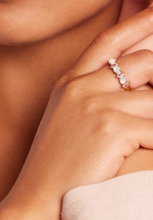 Gouden ring met vijf meerkleurige stenen; ronde en marquise vormen, gladde textuur en een slanke band, gedragen aan een hand met gebruinde huid.