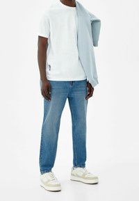 Vit t-shirt med uppvikta ärmar, ljusblå tröja över axeln, blå denimjeans och vita sneakers med ljusa accenter.