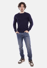 Felpa a maglia blu navy con scollatura tonda, polsini a costine, abbinata a jeans in denim chiaro e sneakers marroni e bianche.