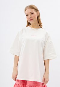 Overgrote witte t-shirt met korte mouwen, voorzien van roze tekst "MIRONS" op de borst. Gepaarde met een roze gelaagde rok. Glad stoftextuur.