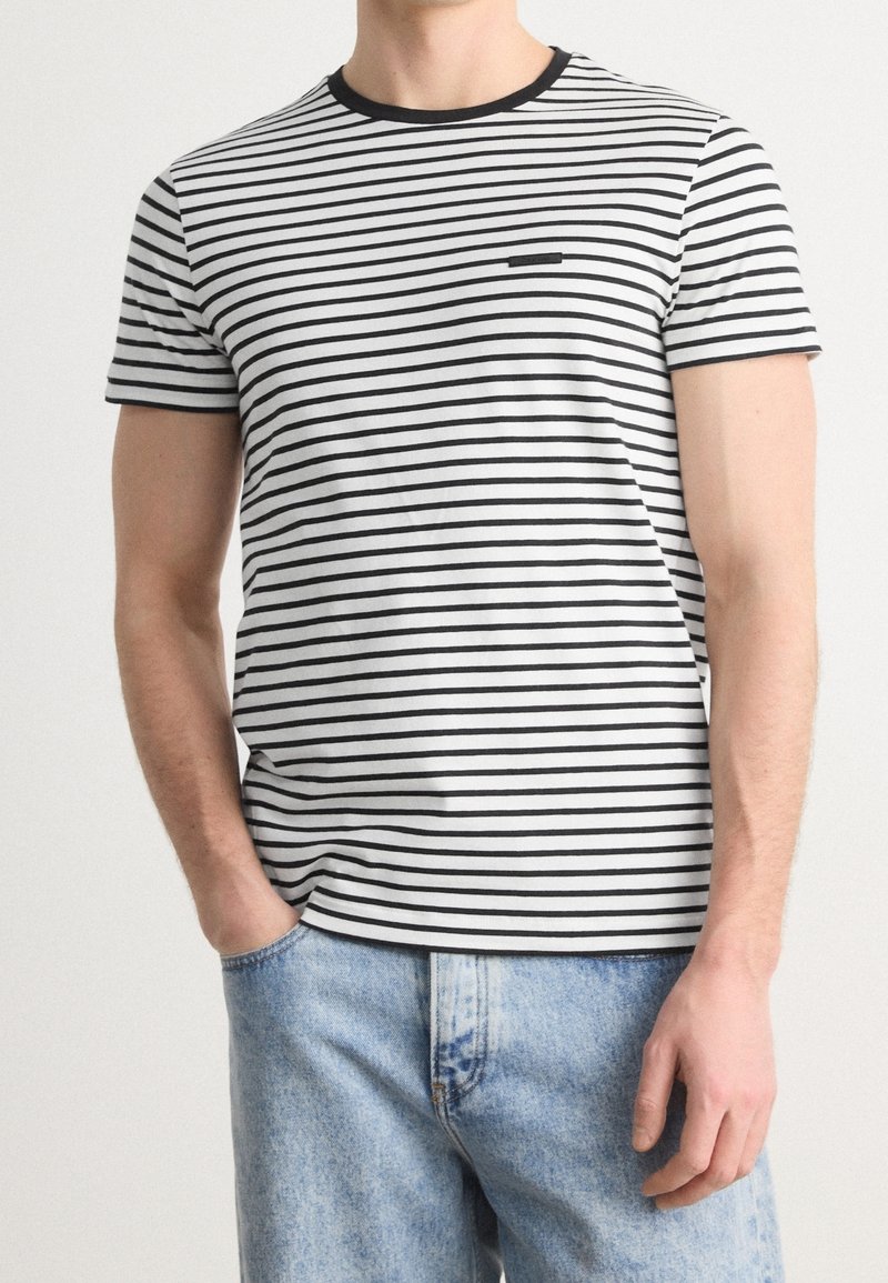 T-shirt à manches courtes en rayures horizontales blanches et noires, avec un col rond noir et un petit logo sur la poitrine. Associé à un jean en denim bleu clair.