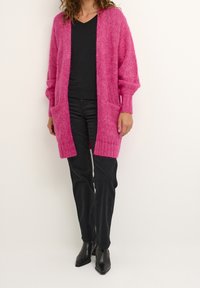 Cardigan lavorato a maglia di un luminoso colore rosa con apertura frontale, orlo a costine e due tasche laterali. Indossato sopra una maglietta nera e abbinato a pantaloni neri e stivali.