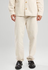 Beige cotton pants with a relaxed fit, featuring side pockets and a smooth texture, paired with black shoes with straps and chunky soles.