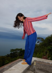 Femme souriante en équilibre sur un étroit rebord en béton à l'extérieur, portant un pull rayé, pantalon bleu et sandales jaunes, avec des arbres et un ciel nuageux en arrière-plan.