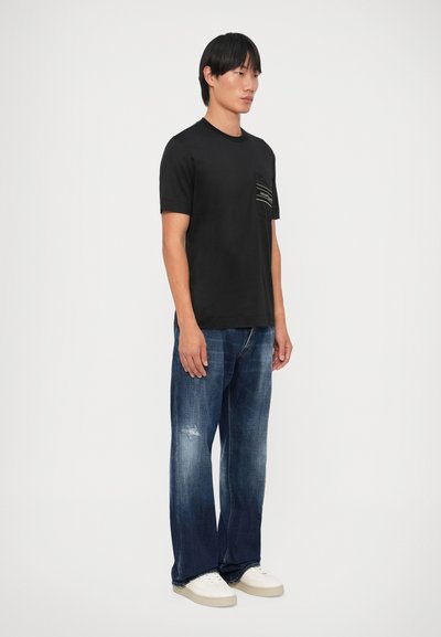 Camiseta de algodão preta com um bolso no peito com riscas horizontais. Usada com jeans azuis desbotados e ténis brancos. Design de corte reto.