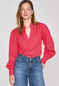 Blusa rossa a fantasia con maniche a sbuffo e scollo a V profondo, abbinata a jeans in denim blu a vita alta. Il tessuto presenta un motivo a quadri.