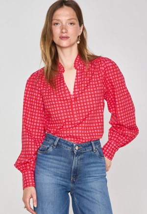 Blouse rouge à motifs avec des manches bouffantes et un décolleté plongeant, associée à un jean en denim bleu taille haute. Le tissu présente un motif à carreaux.