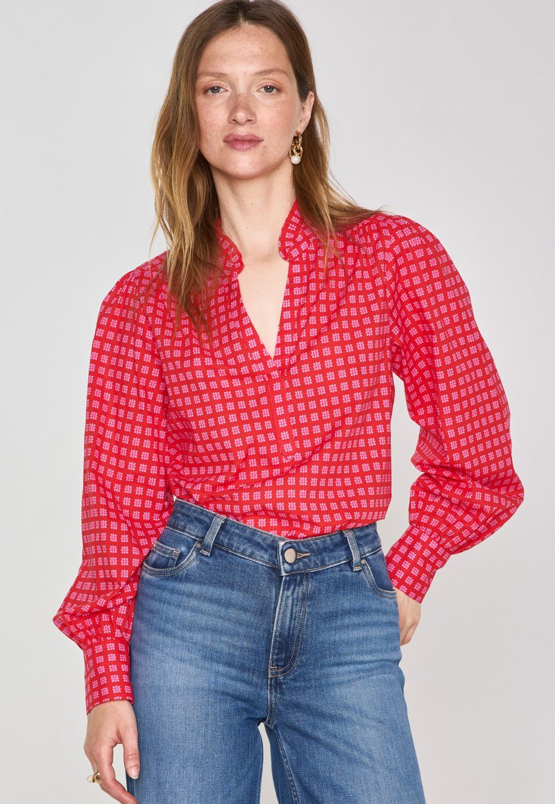 Blusa rossa a fantasia con maniche a sbuffo e scollo a V profondo, abbinata a jeans in denim blu a vita alta. Il tessuto presenta un motivo a quadri.
