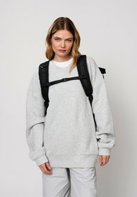 Felpa oversize grigia chiara con orlo e polsini a costine, indossata sopra una maglietta bianca, abbinata a pantaloni grigi e uno zaino nero.
