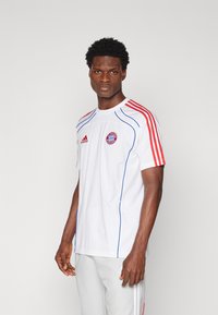 Biała sportowa koszulka z czerwonymi i niebieskimi akcentami. Posiada trzy czerwone paski na ramionach oraz okrągłe logo Bayernu Monachium na piersi.
