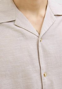 Chemise à manches courtes en lin beige clair, avec un col classique, une texture subtile et un bouton beige au niveau du col.