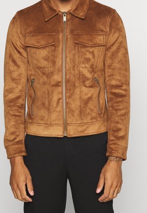 Mężczyzna ubrany w brązową zamszową kurtkę z przednim zamkiem błyskawicznym, dwoma kieszeniami na klapki na piersi oraz dwoma zamykanymi na zamek kieszeniami po bokach, w połączeniu z czarnymi spodniami.