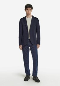 Marinblazer med snäpplabb, två framfickor, över en ljusgrå t-shirt, ihop med mörka jeans och svarta skor.