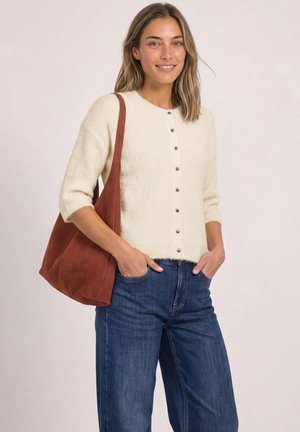 Femme portant un pull crème boutonné et un jean bleu, portant un grand sac à bandoulière marron, mains dans les poches et souriante.