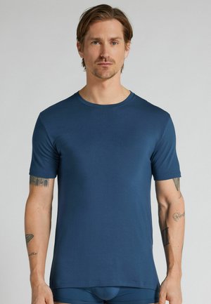 Mężczyzna z krótkimi brązowymi włosami, ubrany w gładki niebieski t-shirt z krótkim rękawem, stojący na jednolitym jasnoszarym tle.