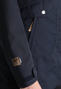 Manche de veste bleu marine foncé avec un logo patch marron, sangle de poignet ajustable avec Velcro et une poche zippée avec une petite languette en cuir.