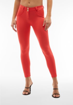 Leggings rossi aderenti con una texture liscia, dotati di una cintura con bottoni e due tasche frontali. Indossati con sandali neri con cinturini.