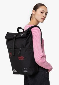 Svart rull-top ryggsäck med justerbara remmar, framficka, röda grafiska inslag och en hållbar tygstruktur.