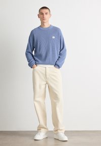 Sweater azul claro com nervuras e decote redondo, usado sobre uma camisa branca, combinado com calças retas em creme e ténis brancos.