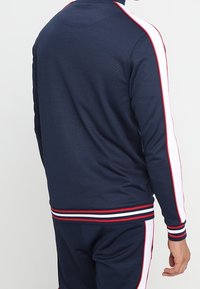 Chaqueta deportiva de color azul marino con acentos a rayas rojas y blancas en las mangas y el cuello. Presenta una cintura acanalada y un tejido texturizado.