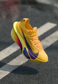 Gelbe gestrickte Sneaker mit einer farbverlaufenden Sohle von Lila zu Orange, strukturiertem Mesh-Obermaterial und einem auffälligen Logo in Lila an der Seite.
