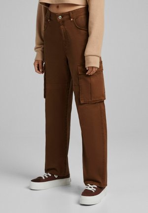 Bruine cargobroek met een losse pasvorm, voorzien van grote zijzakken, een knoopsluiting en contrasterende stikseldetails. Gepaard met donkere sneakers.