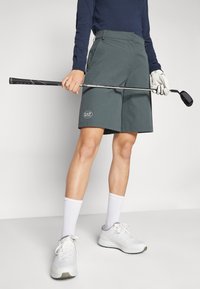 Tamsiai žalios golfu šortai turi lygų paviršių ir logotipo etiketes. Su baltais sportiniais batais ir kojinėmis. Golfų lazdą laiko dešinėje rankoje.