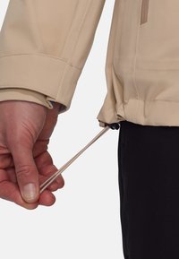 Helles beiges Jacket mit Zugband am Bündchen. Der Stoff wirkt glatt und wird mit schwarzen Unterteilen kombiniert.