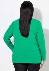 Zielona bluzka z długim rękawem o gładkiej fakturze, zaokrąglonym brzegu i luźnym kroju, zestawiona z ciemnymi jeansami oraz prostymi czarnymi włosami widocznymi z tyłu.