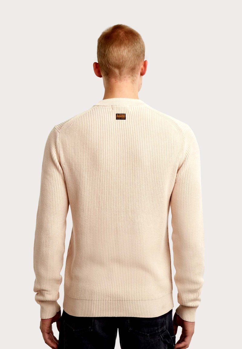 Suéter de punto acanalado en beige claro con mangas largas, cuello redondo y una pequeña etiqueta de marca en la parte trasera. Textura sutil en toda la prenda.