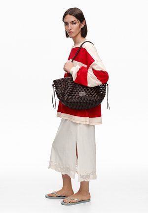 Kobieta w czerwono-białym oversize swetrze, białej midi spódnicy z rozcięciem, szarych japonkach, niosąca dużą ciemną plecioną torebkę na ramieniu.