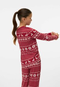 Dziewczyna z brązowym kucykiem, ubrana w długą bluzkę z czerwono-białym wzorem reniferów i śnieżynkami oraz spodnie, wyciągająca ramiona do przodu.