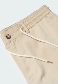 Taille de pantalon beige avec élastique, fermeture par bouton, passants de ceinture et cordon blanc avec embout métallique.