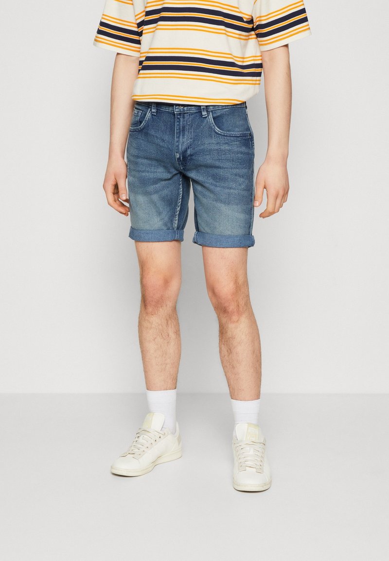 Shorts en denim bleu avec des revers, présentant un design classique à cinq poches. Portés avec une chemise à rayures à manches courtes et des baskets blanches.