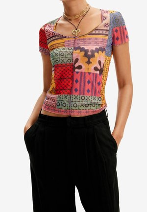 Žena má na sobě přiléhavý krátký rukáv s patchworkovým vzorem, černé kalhoty s vysokým pasem a vrstvené zlaté náhrdelníky.