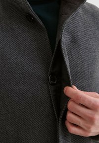 Grauer Fischgrätmantel mit hohem Kragen, ausgestattet mit einem schwarzen Knopfverschluss und einem glatten Futter. Eine Hand richtet das Revers in der Nähe des Knopfs.